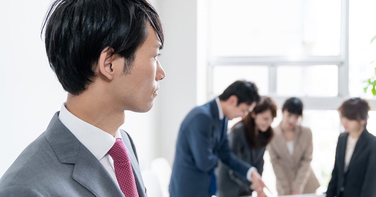 職場で同僚たちの様子を見るスーツ姿の男性