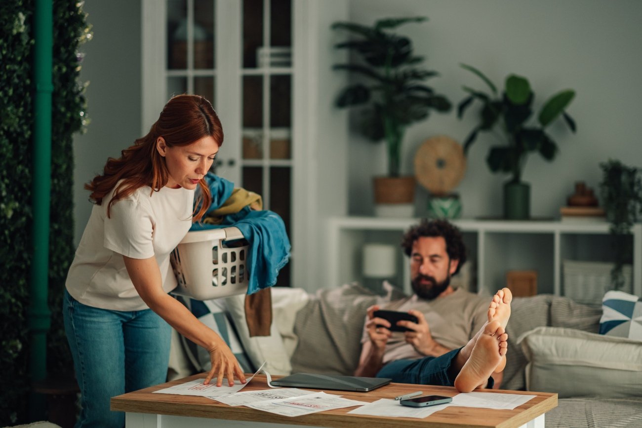 Woman managing household chores and finances, while partner relaxes, highlighting unequal division of labor
