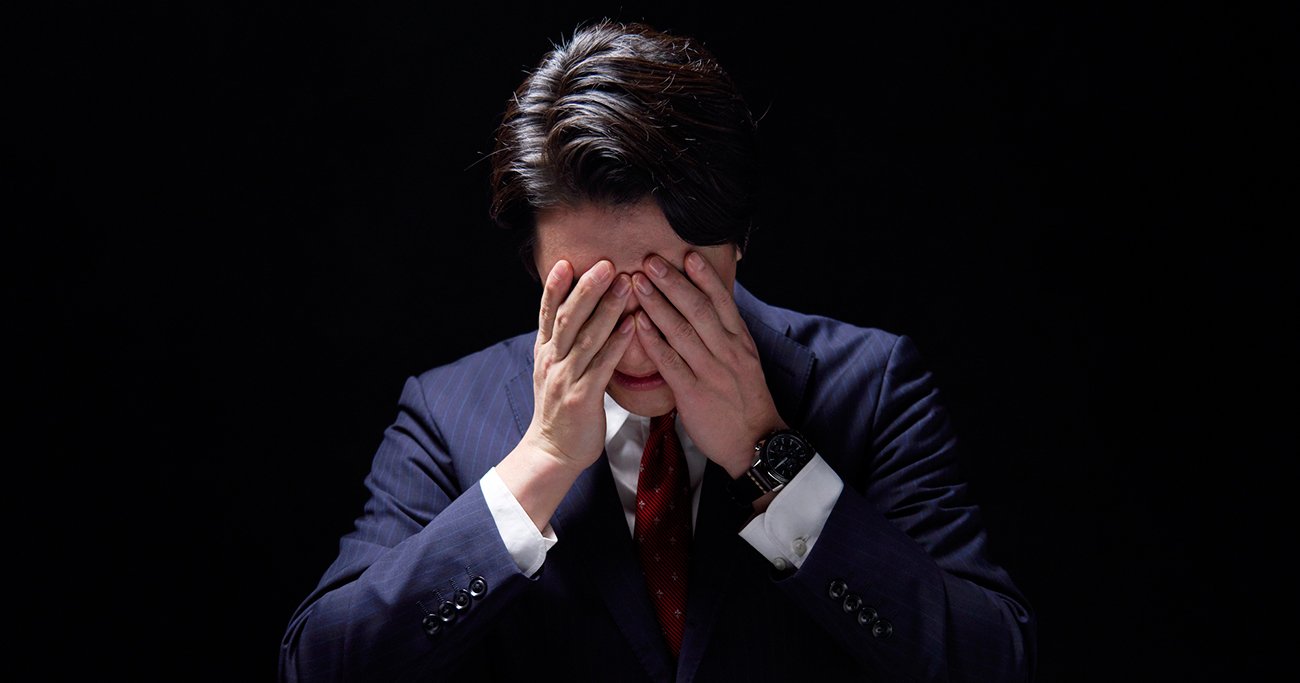 顔を手で覆いうつむくスーツ姿の男性