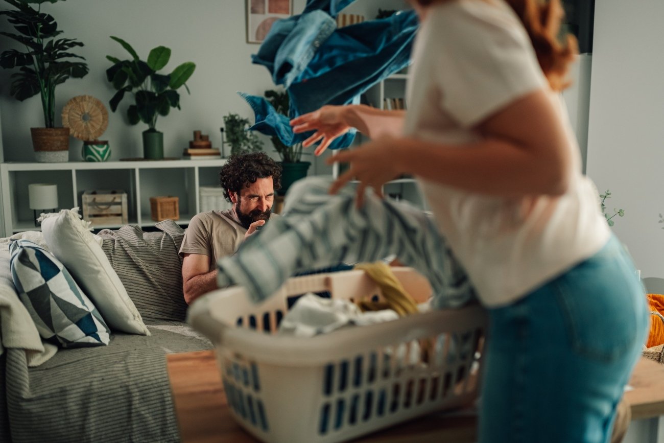 Couple arguing about housework at home, wife throwing dirty laundry from basket at lazy husband
