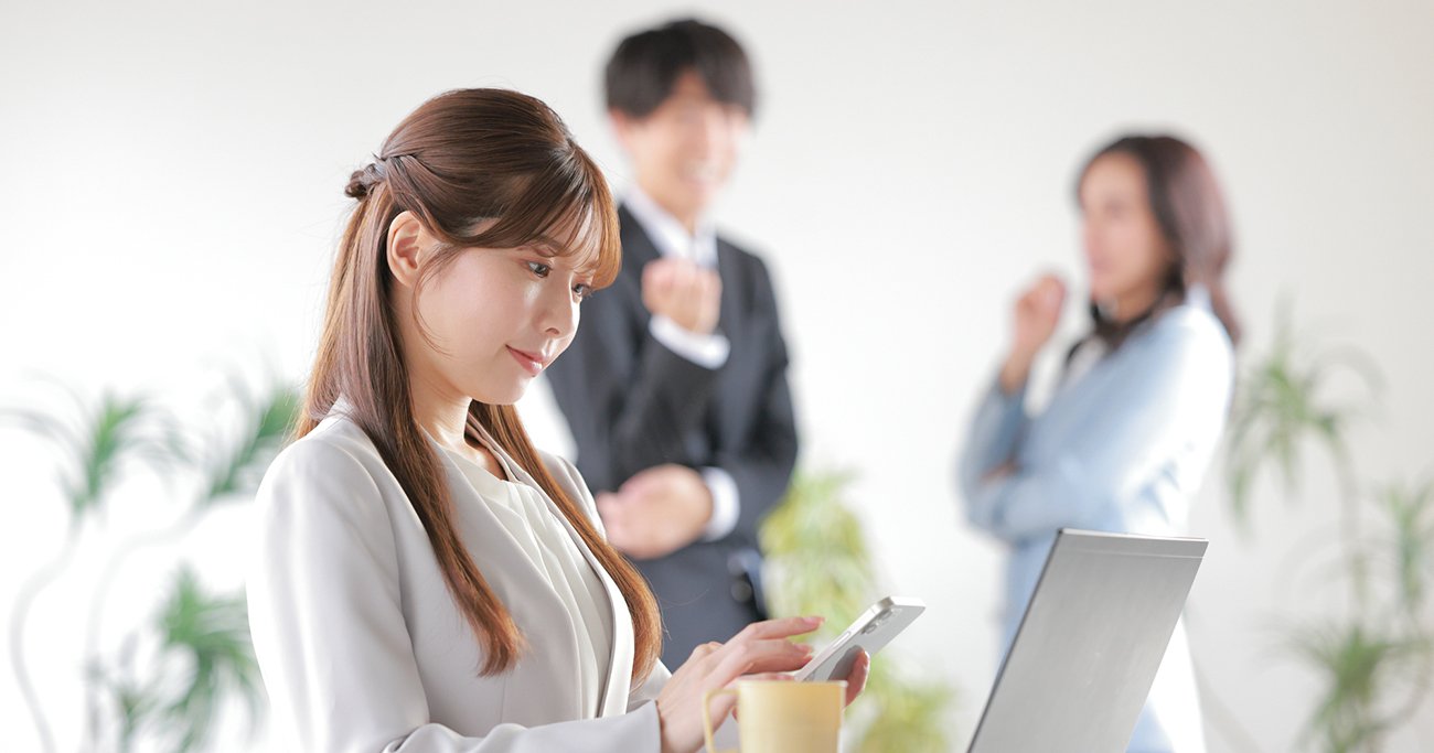 スマホを見る女性社員の背後で、同僚たちが会話をする様子