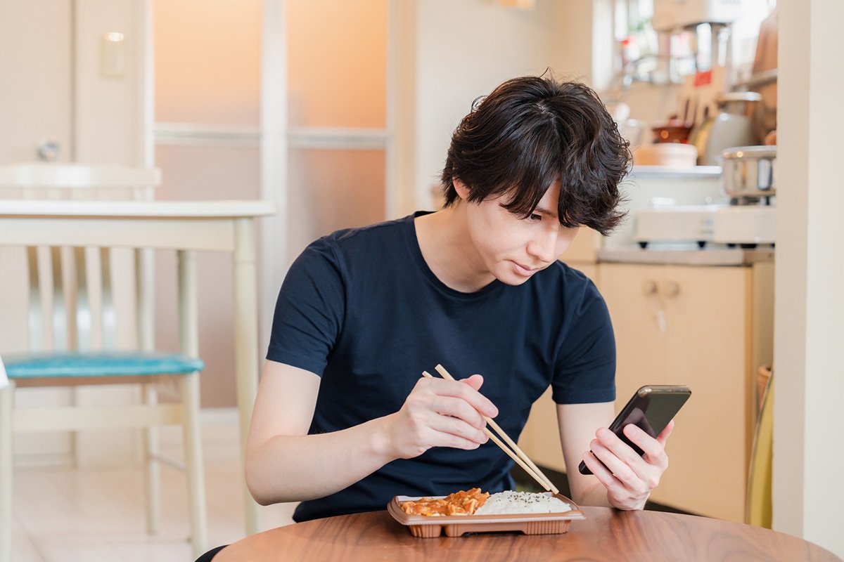 コンビニ弁当を食べながらスマホを見る男性